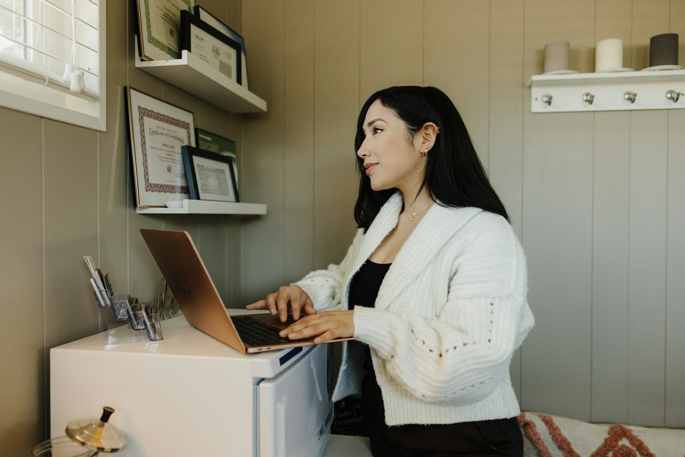 A woman types on a laptop at a small home desk with framed certificates on the wall