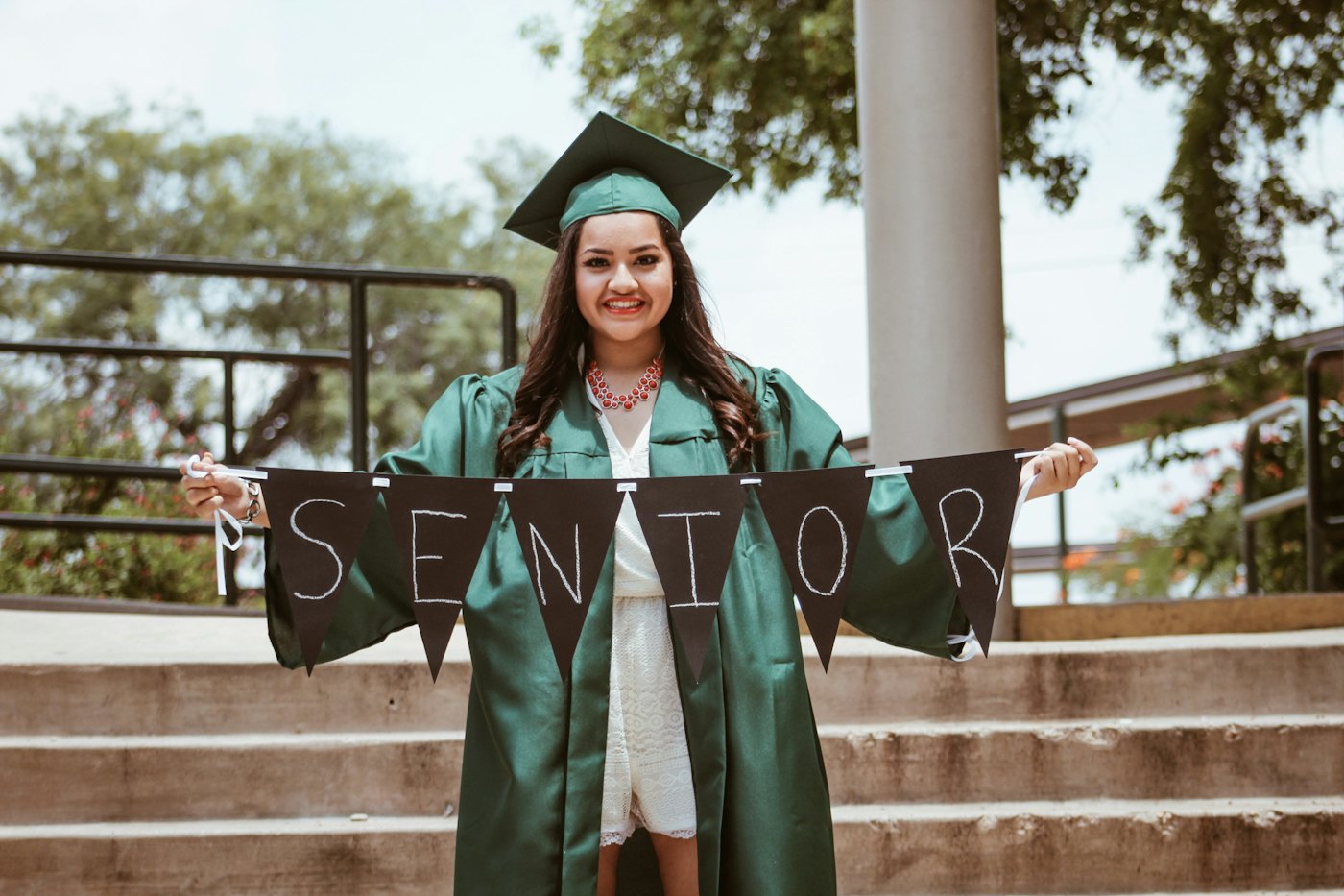 A smiling graduate in a green cap and gown holds a SENIOR banner