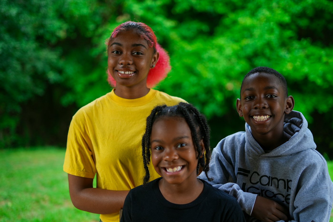 Three smiling kids, a teen girl, a younger girl and a boy, stand together outdoors