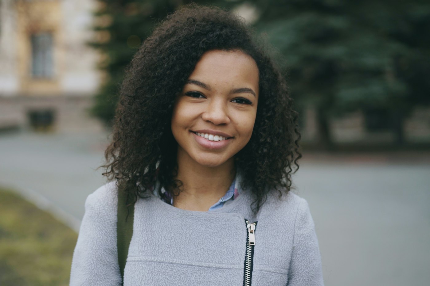 A smiling teen girl with curly hair and a backpack stands outdoors