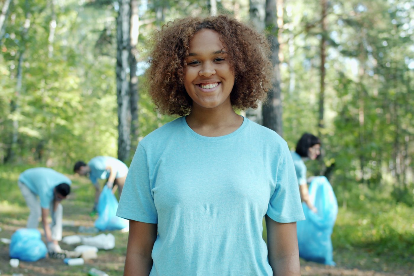 A smiling young volunteer in a blue T-shirt stands in a wooded park while others collect litter behind her
