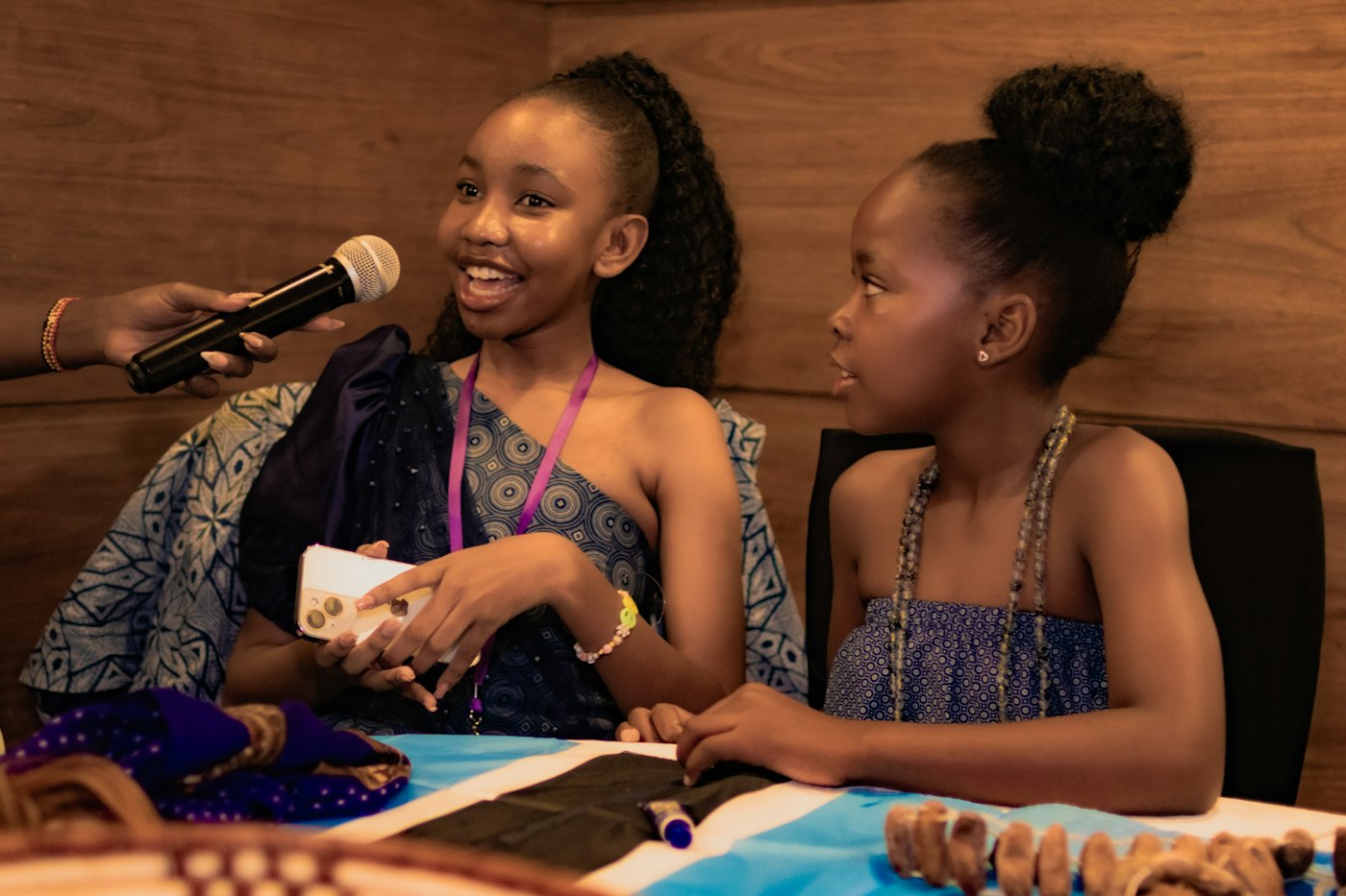 A smiling girl speaks into a microphone at a table displaying handmade baskets and beadwork, with a younger girl beside her