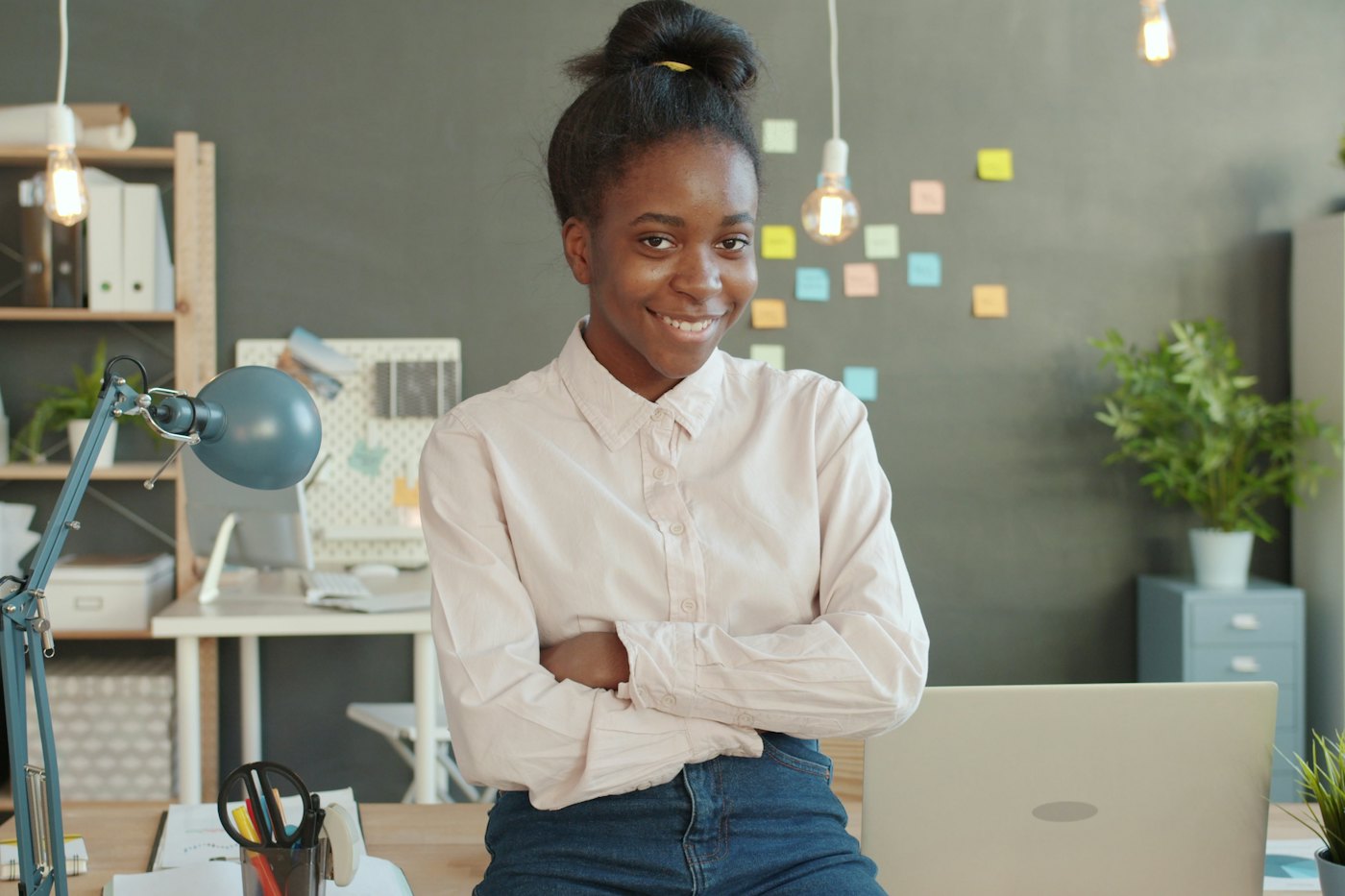 A young woman in a button-down shirt smiles with arms crossed, leaning on a desk in a bright office